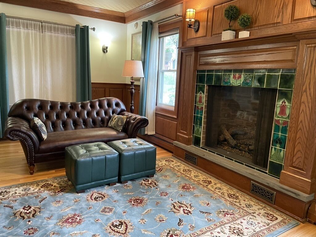 A living room with a brown leather sofa, a matching ottoman, a patterned rug, a fireplace with green tiles, and a wooden mantel.