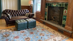 A living room with a brown leather sofa, a matching ottoman, a patterned rug, a fireplace with green tiles, and a wooden mantel.