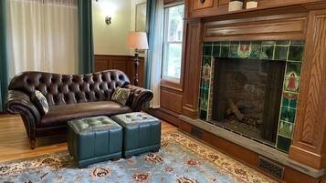 A living room with a brown leather sofa, a matching ottoman, a patterned rug, a fireplace with green tiles, and a wooden mantel.