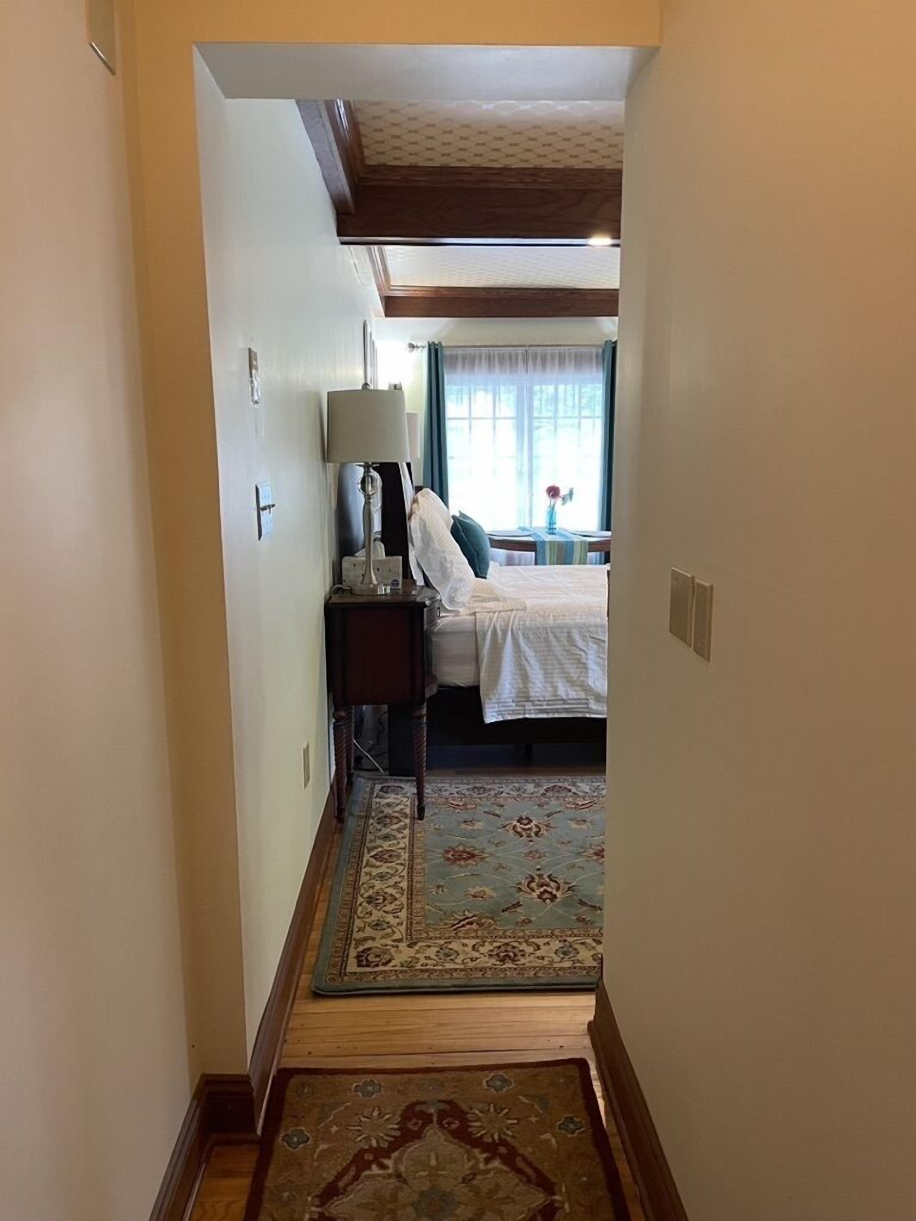 Un corridoio stretto che conduce a una camera da letto con un letto, un comodino, una lampada e una finestra con tende.