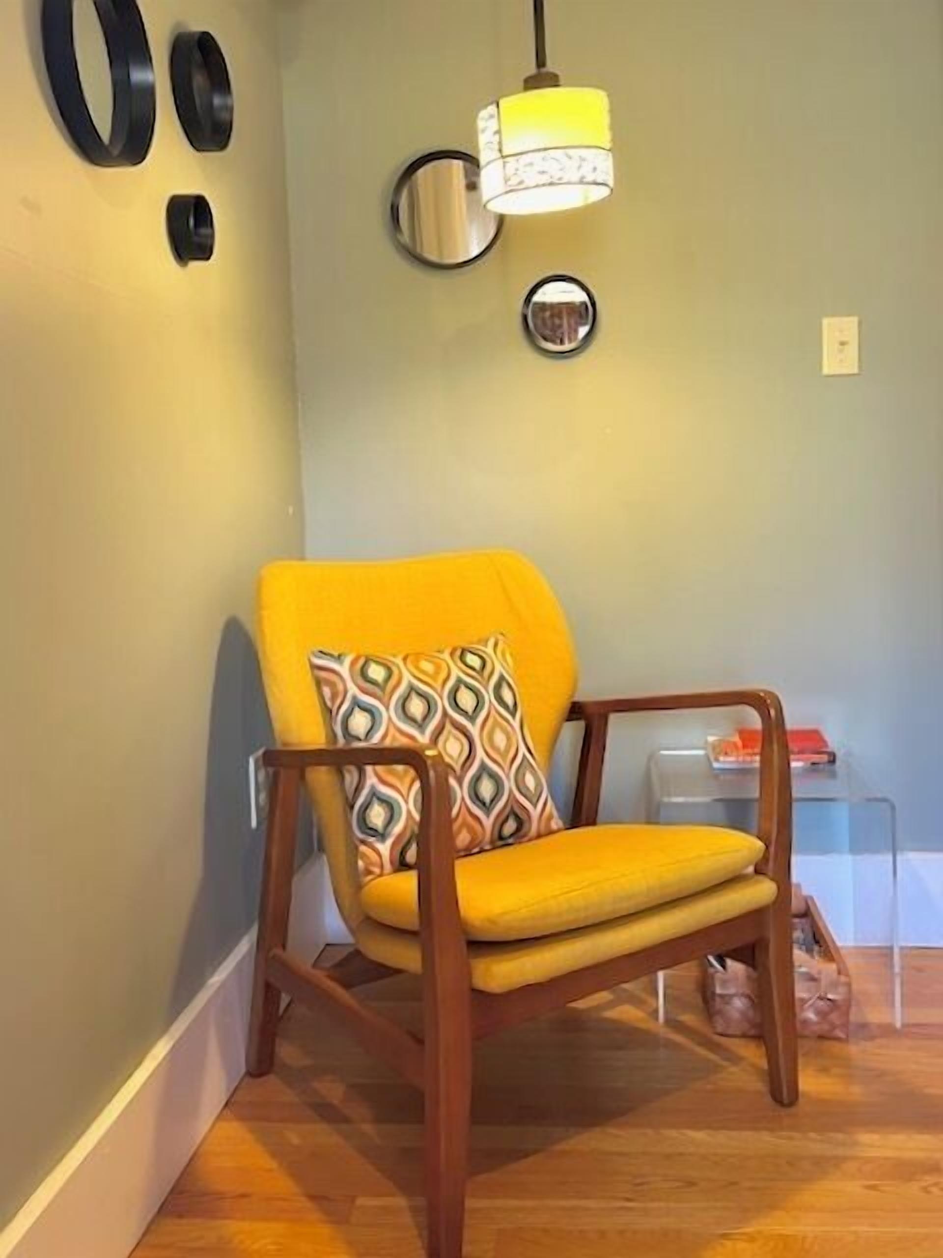 A corner room with a wooden chair, a yellow cushion, and a wall-mounted lamp.