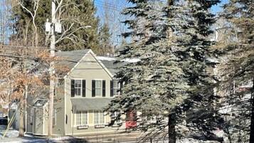 Une maison à deux étages, dotée d’un toit à pignon, entourée d’arbres et recouverte de neige.