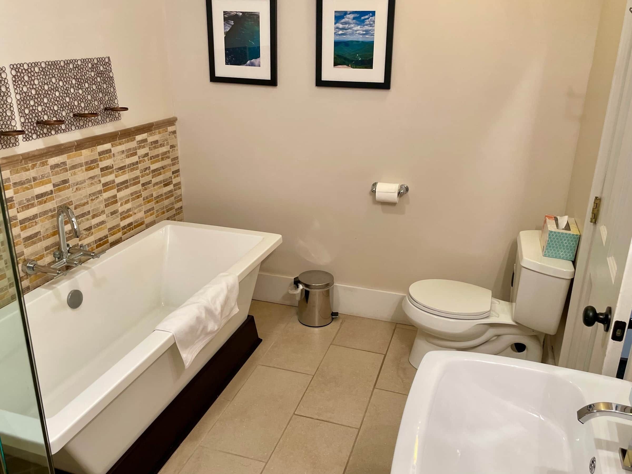 Een moderne badkamer met een vrijstaand bad, toilet en wastafel. Twee ingelijste foto's hangen aan de muur.