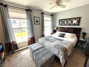 Un dormitorio con cama, un banco, ventilador de techo y ventana con cortinas.