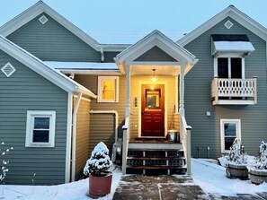Una casa de dos pisos con una puerta roja, un pequeño porche y un paisaje cubierto de nieve.