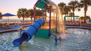 A water play area with a slide and water jets, surrounded by palm trees and beach chairs.