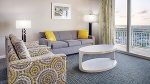 A modern hotel room with a grey sofa, patterned armchair, and a round coffee table. There are two lamps, a framed artwork on the wall, and a large window with a view of the city.