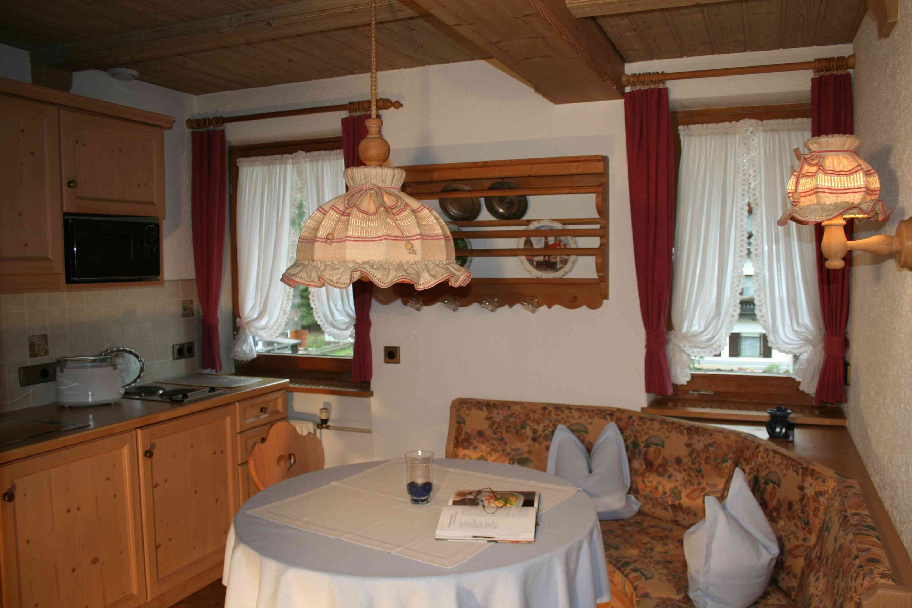 Ein gemütliches Zimmer mit Holzbalkendecke, einem Esstisch mit weißem Tischlauf, einer Couch mit Kissen und einem Fenster mit Vorhängen.