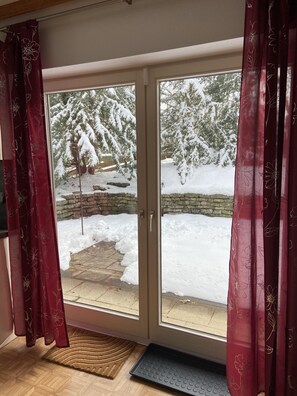 Een kamer met een schuifdeur die uitkomt op een besneeuwde omgeving, met een stenen muur en een houten terras.