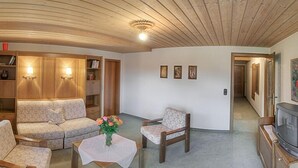 Una sala de estar acogedora con un sofá, sillones, una mesita con flores, un televisor y estanterías de madera con luces de pared.