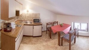 Cocina compacta con microondas, pileta y armarios de madera. Se ve una mesa de comedor con sillas y mantel rojo.