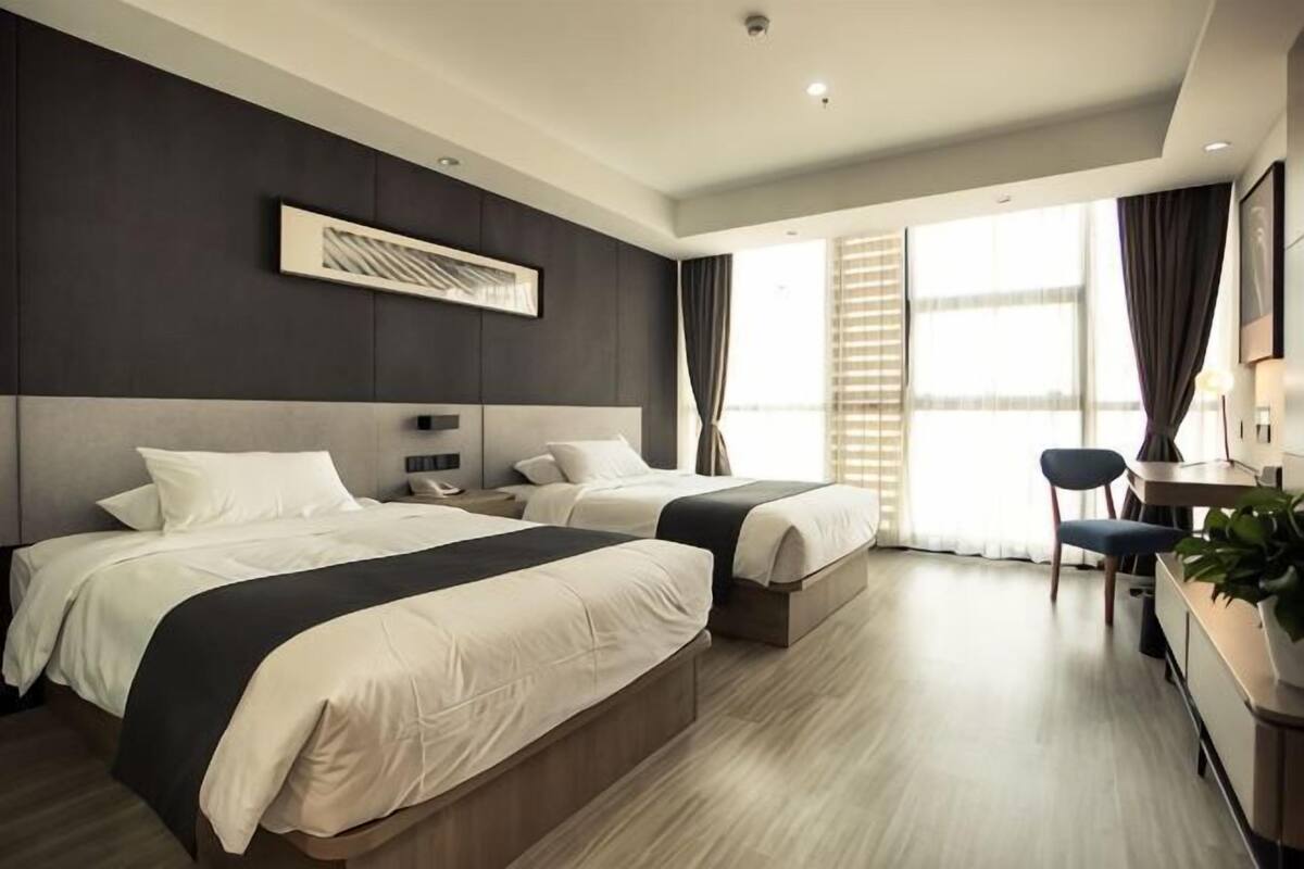 Habitación de hotel con dos camas, un escritorio, una silla y un gran ventanal con cortinas.