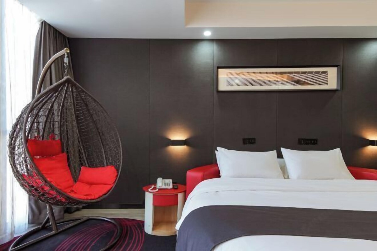Una habitación de hotel moderna con una silla colgante, una cama con ropa blanca, una butaca roja, una mesita de noche pequeña con un teléfono y un cuadro en la pared.
