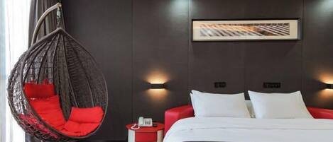 Una habitación de hotel moderna con una silla colgante, una cama con ropa blanca, una butaca roja, una mesita de noche pequeña con un teléfono y un cuadro en la pared.