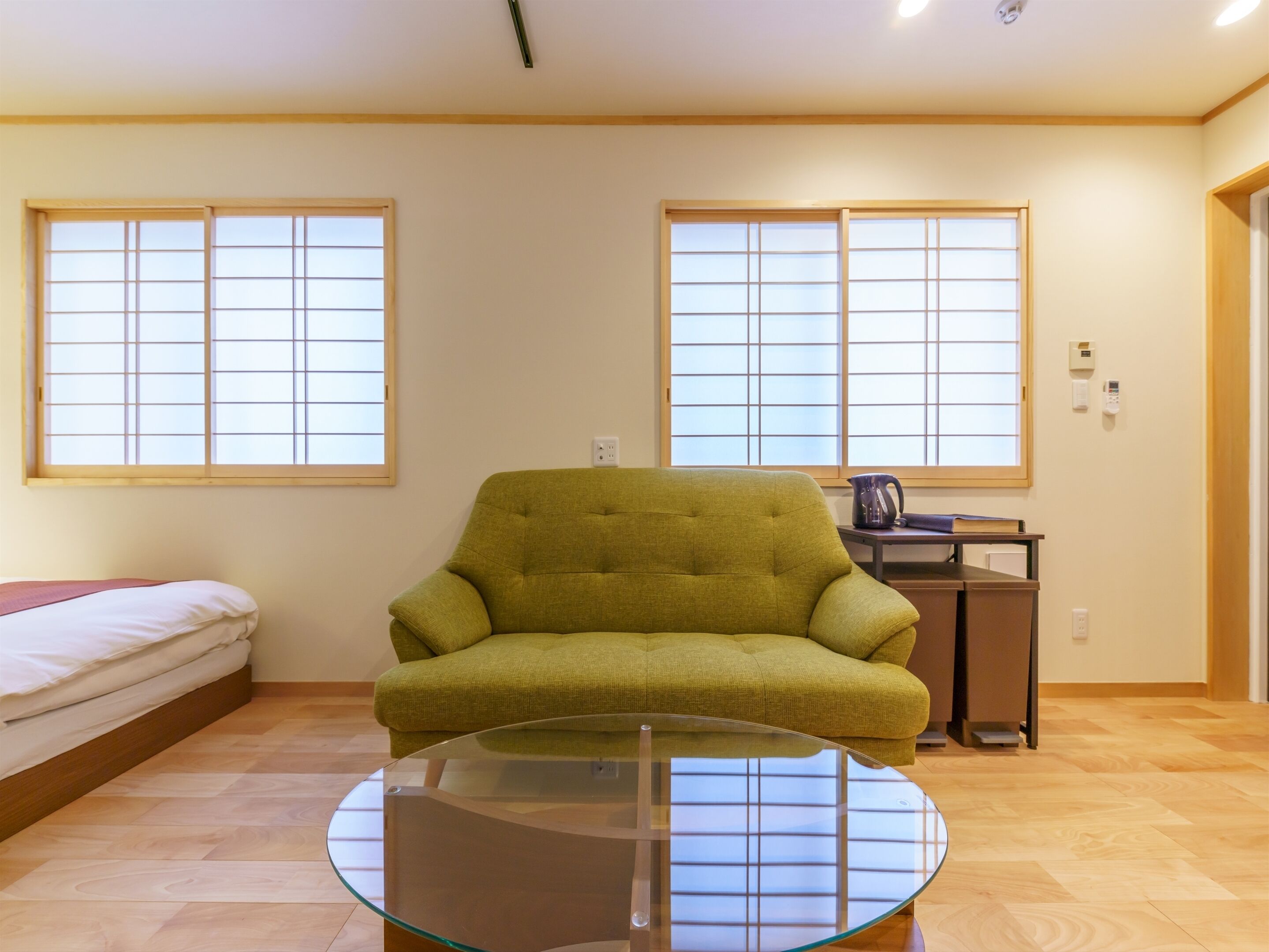 A room with a green sofa, a glass coffee table, a bed with a white mattress, and two windows with wooden frames.