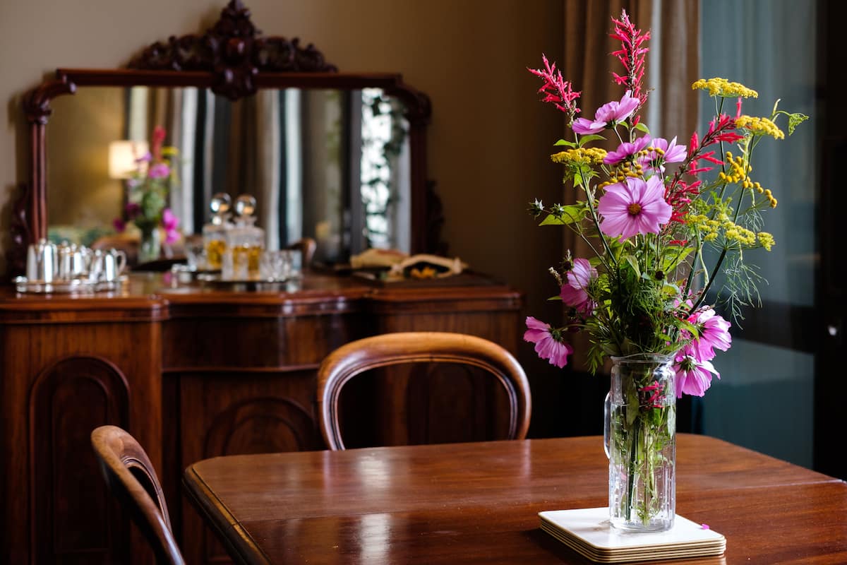 A wooden dining table with a vase of flowers on a coaster, a wooden chair, and a mirror on the wall.