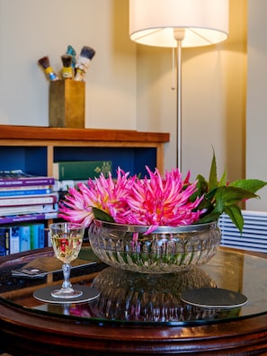 A glass bowl with pink flowers on a reflective surface, a wine glass, and a lamp in the background. - Bell Hill Apartments (Dunedin)