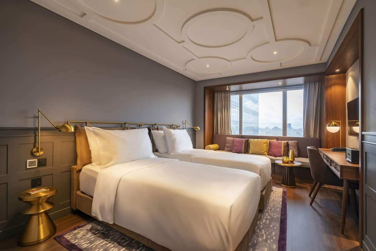A hotel room with two beds, a desk, and a view of a cityscape.