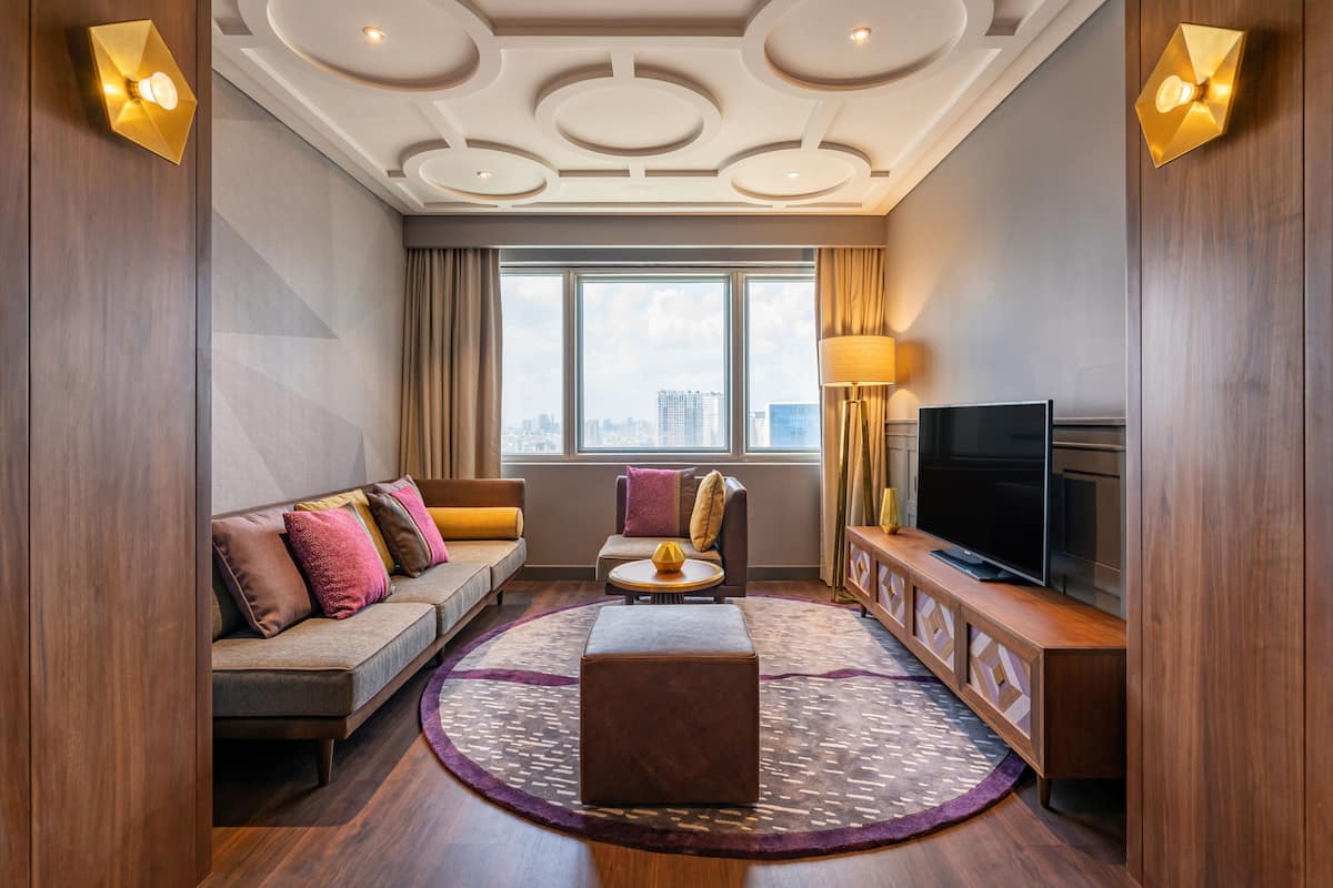 A modern hotel room with a sofa, armchair, ottoman, TV, and a large window offering a city view.