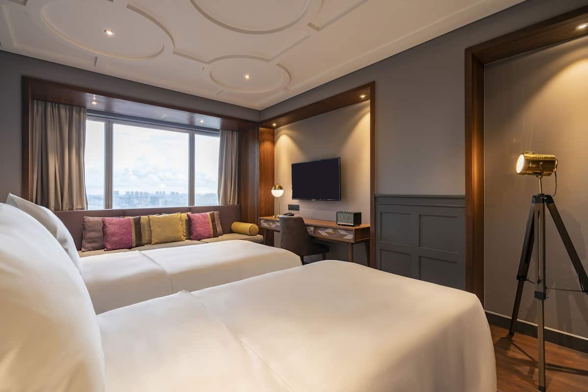 A hotel room with two beds, a desk with a computer, a TV, and a window with a city view.