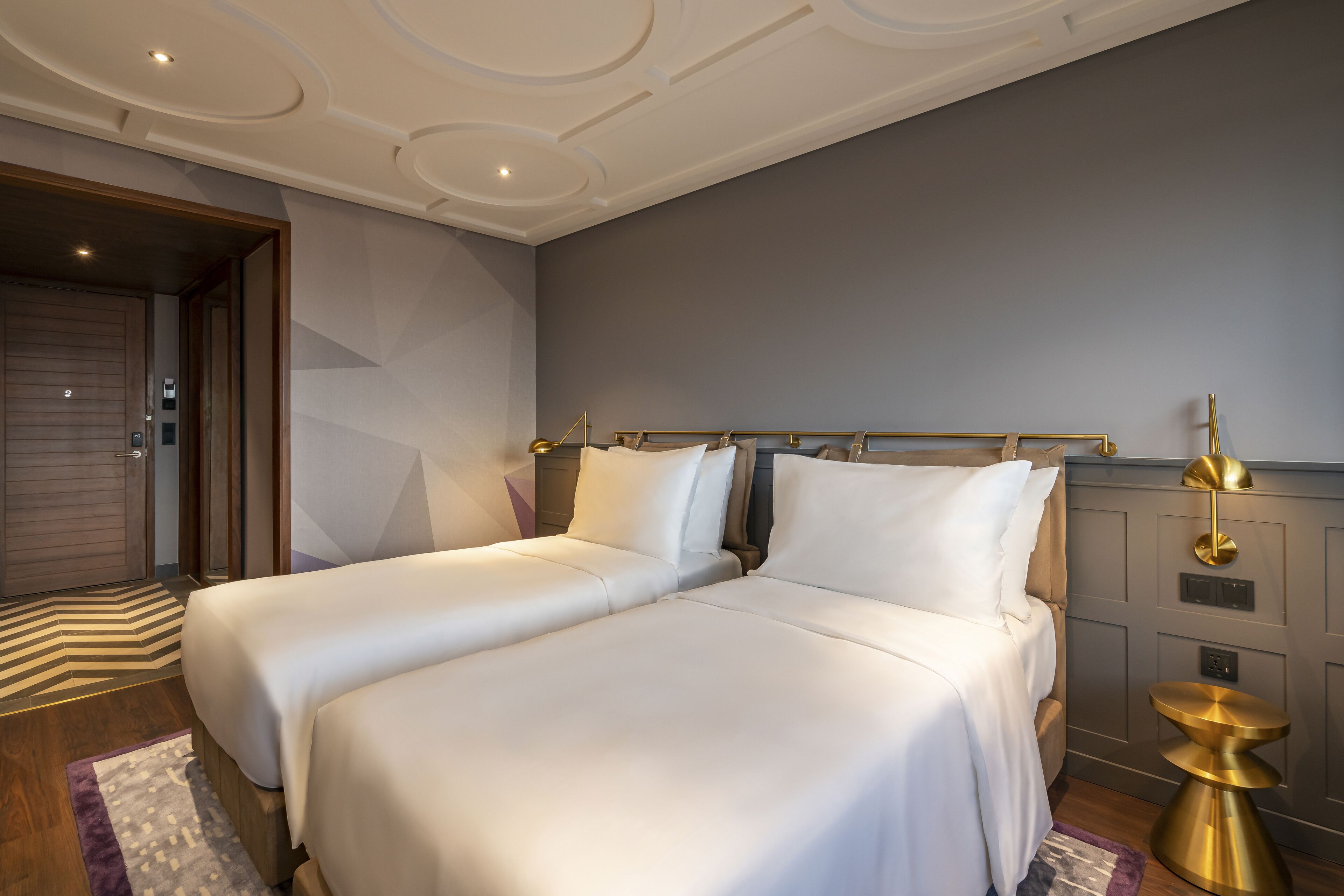 A modern hotel room with two beds, a wooden door, a patterned rug, and a gold accent on the nightstand.