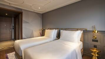 A modern hotel room with two beds, a wooden door, a patterned rug, and a gold accent on the nightstand.