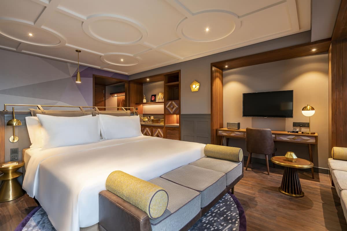 A modern hotel room with a large bed, a desk, a TV, and a seating area.