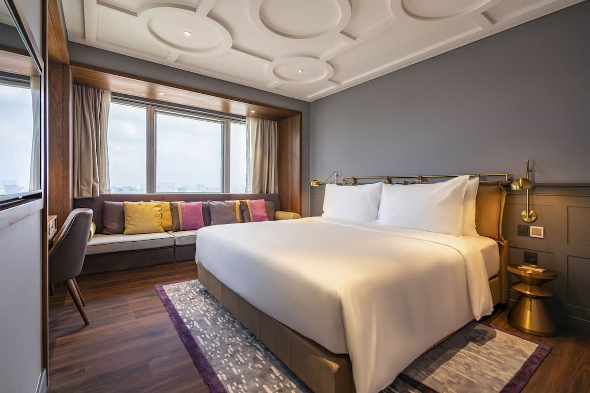 A modern hotel room with a large bed, a seating area with cushions, a view of the city, and a stylish ceiling design.