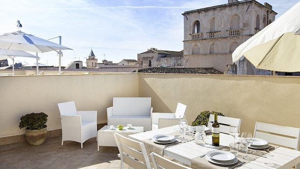 An outdoor patio with white wicker furniture, a table set for dining, and a view of historic buildings in the background. - Appartamento Anice A: A welcoming apartment situated in a central location, at a short distance from the main tourist attractions of the city, with Free WI-FI. (Trapani)