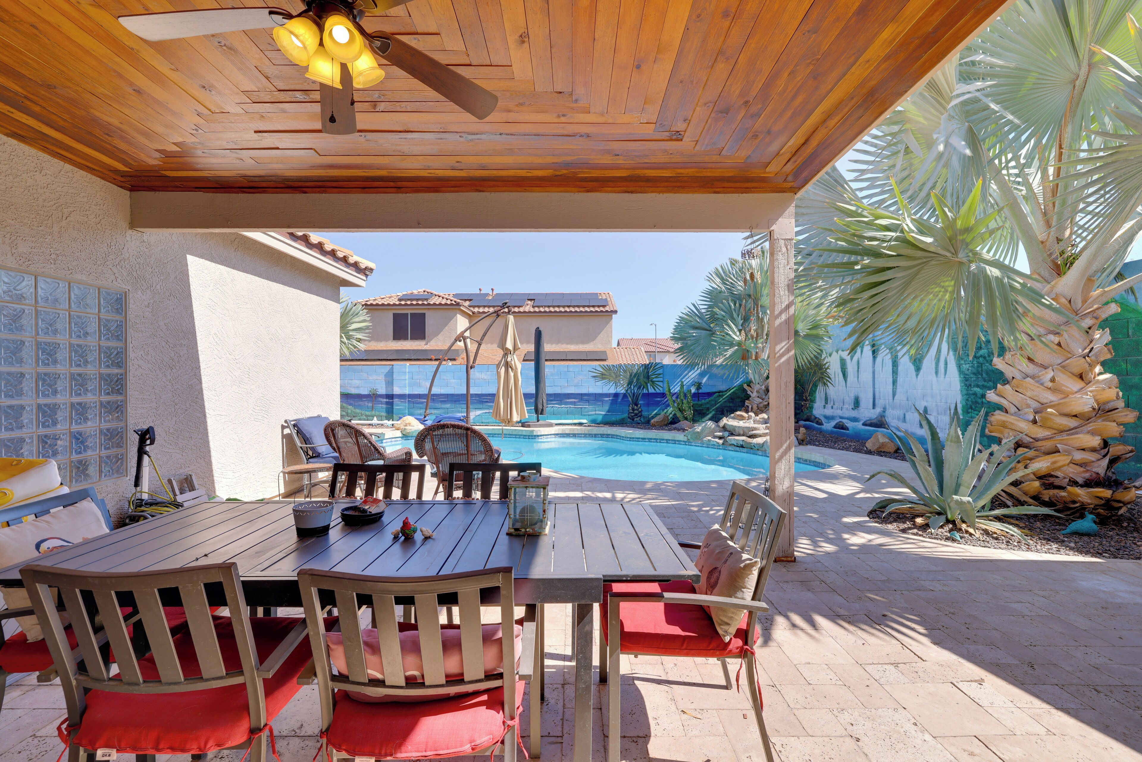 An outdoor patio with a wooden table and chairs, a ceiling fan, and a view of a swimming pool and a house in the background.