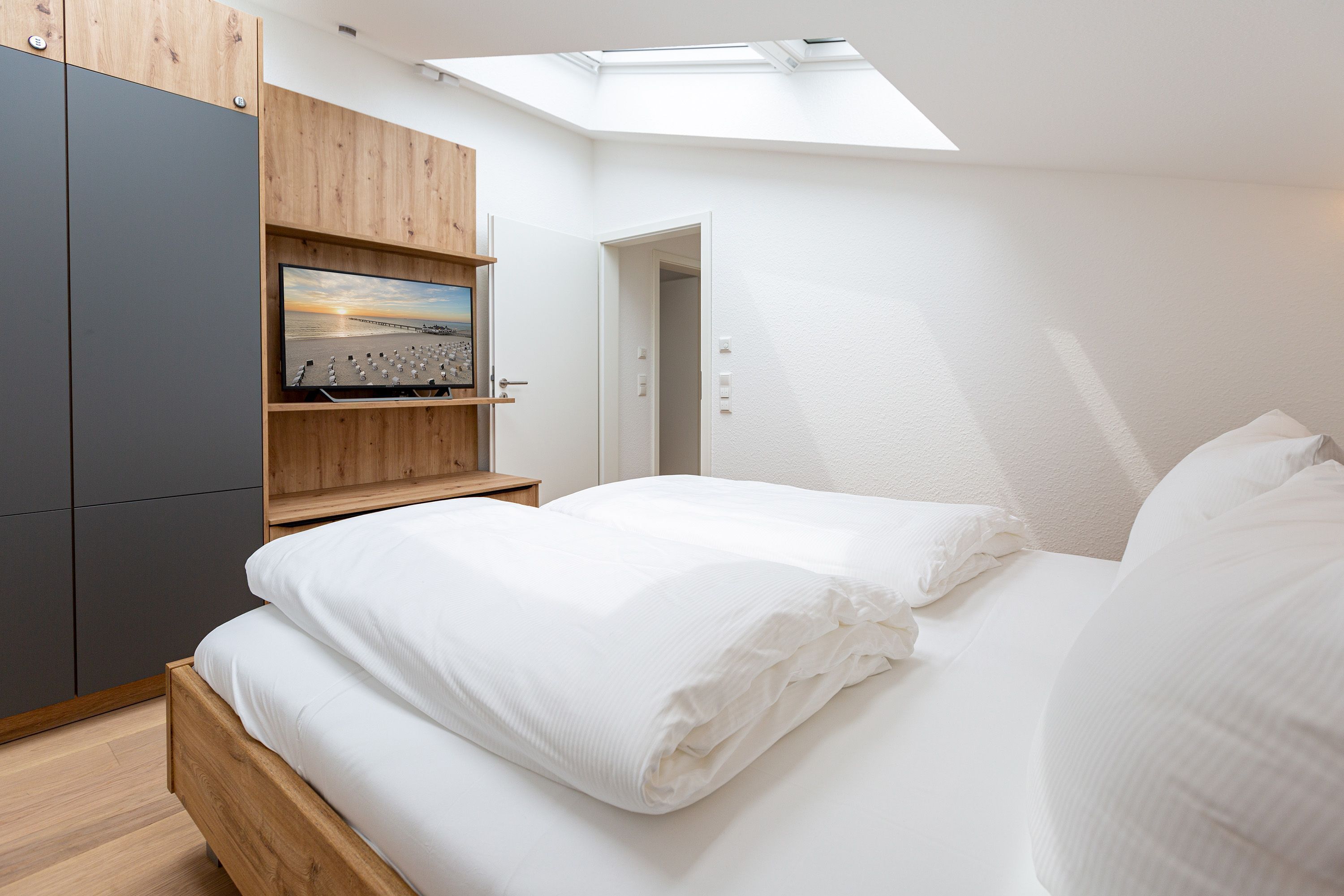 A bedroom with a skylight, a bed with white linens, a wooden wardrobe, and a flat-screen TV on a wooden shelf.