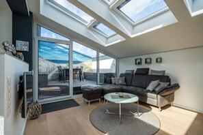 Un salon moderne doté d’un grand canapé d’angle, d’une porte vitrée donnant sur une terrasse extérieure et de fenêtres de toit.