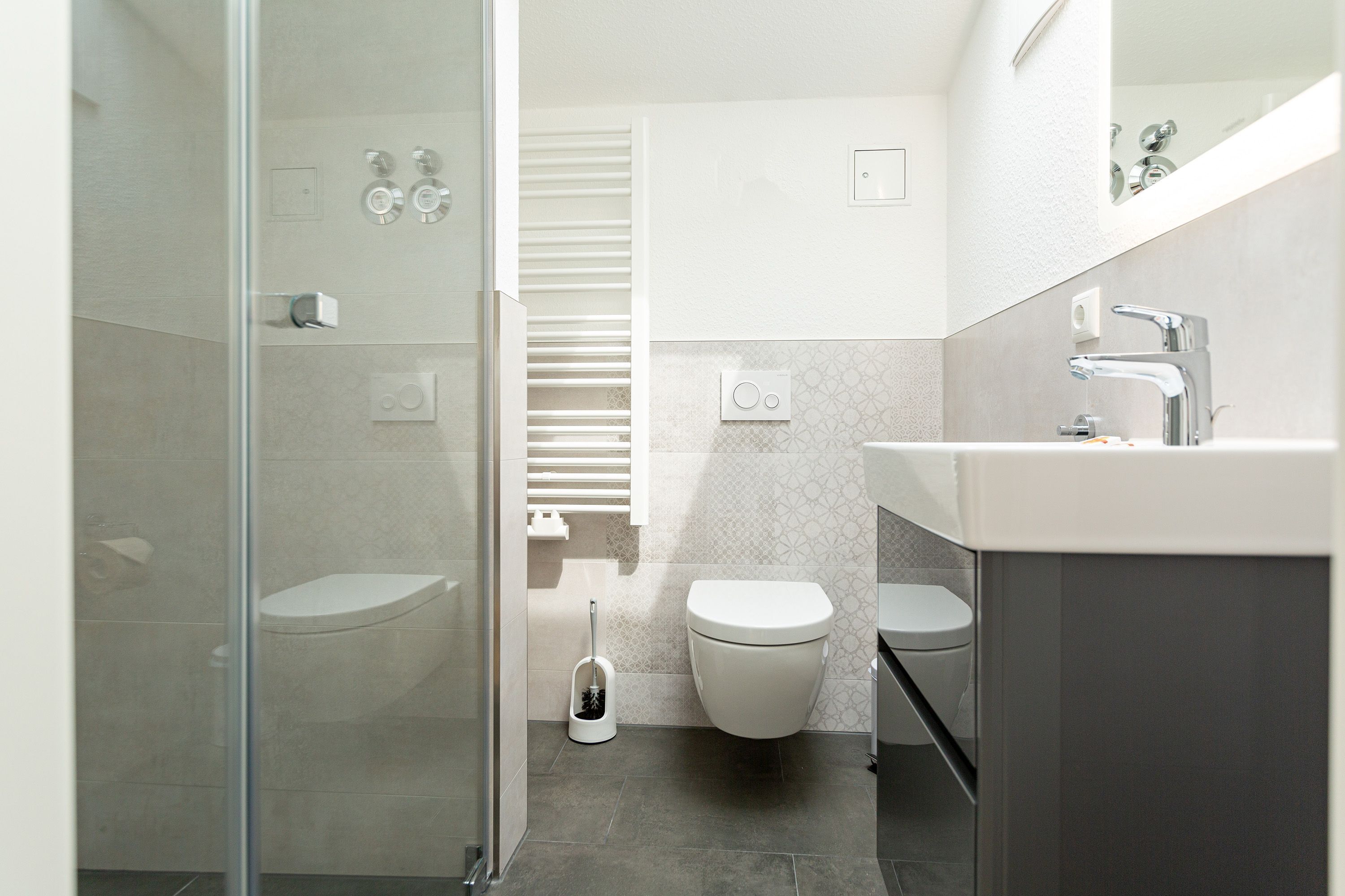 A modern bathroom with a glass shower enclosure, two toilets, a sink, and a toilet brush.