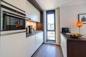 Une cuisine moderne équipée d’un four encastré, d’une bouilloire et d’un four à micro-ondes. On y trouve une fenêtre avec des rideaux, une petite table avec des fruits et un tableau encadré au mur.
