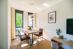 Un coin repas comprenant une table, des chaises, un luminaire suspendu, un tableau encadré et une fenêtre avec des rideaux.