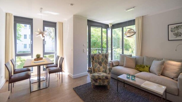 Ein modernes Wohnzimmer mit Essbereich, großen Fenstern, einer Couch, einem Couchtisch und einem gemusterten Sessel.