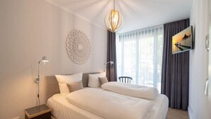 Ein Hotelzimmer mit Bett, Nachttisch, Wandlampe, Wanddeko und einem Fenster mit Vorhängen.