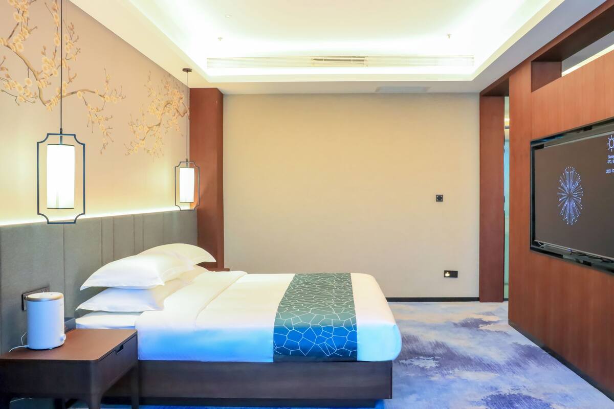 A modern hotel room with a bed, nightstand, wall-mounted TV, and decorative wall art.