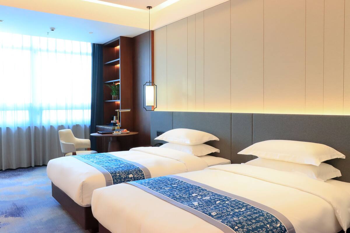 A hotel room with two beds, a desk, a chair, and a bookshelf.