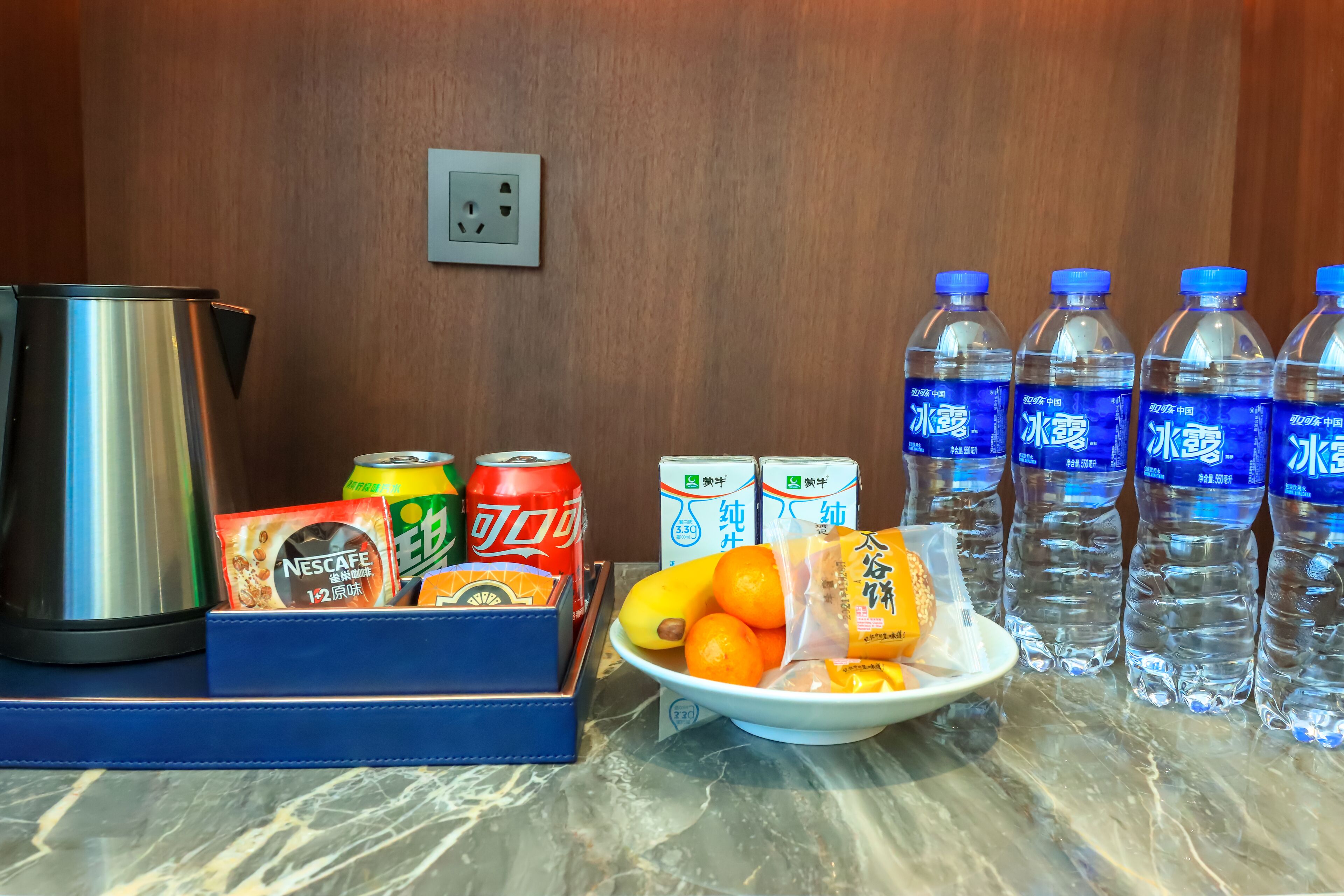A breakfast setup with a kettle, Nescafe, orange juice, water bottles, and fruits on a marble table.