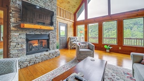 A cozy living room with a stone fireplace, wooden floors, and large windows offering a view of the outdoors. - Decks & Mountain Views: Waynesville Getaway! (Waynesville)