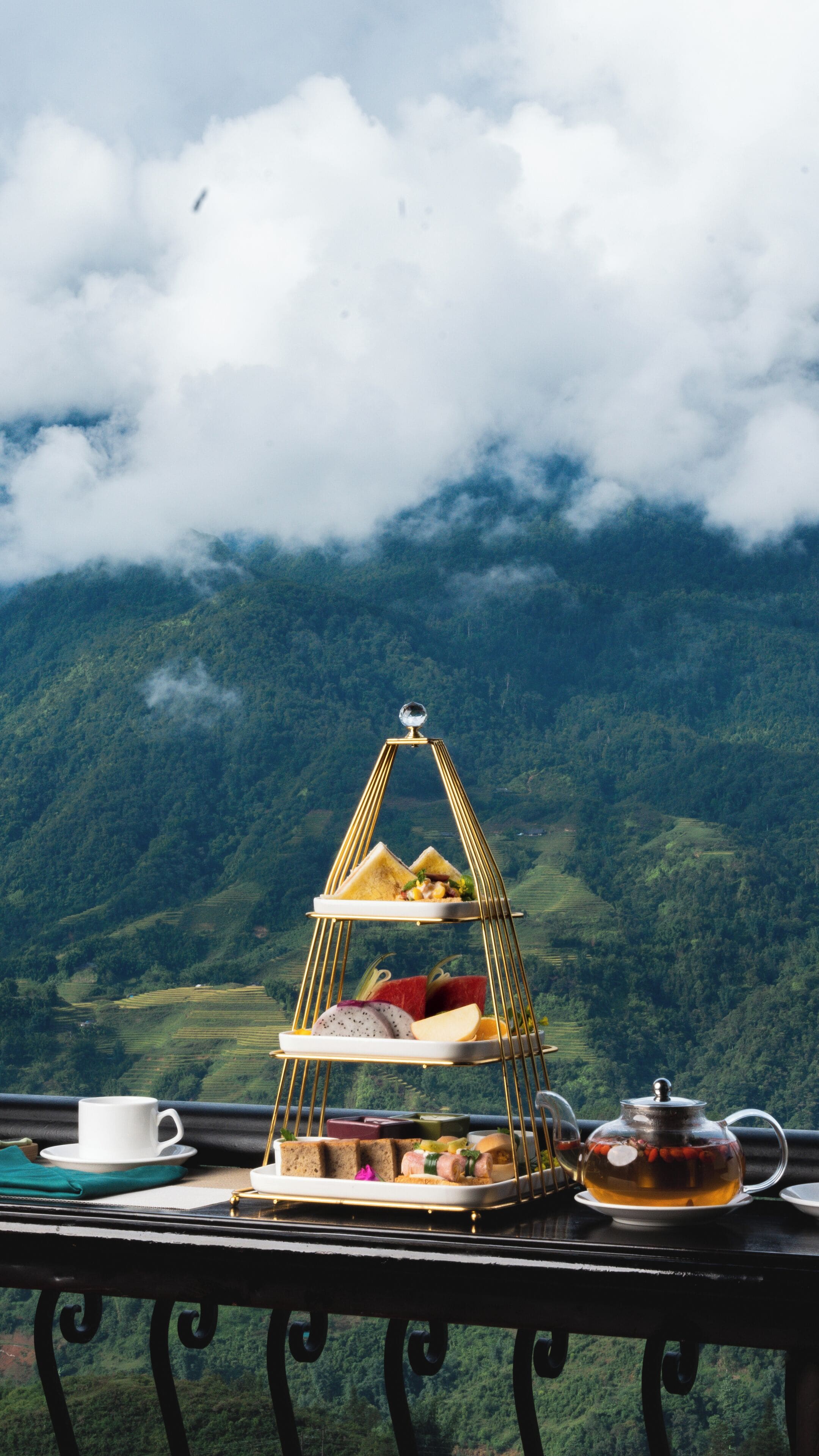 A three-tiered tray with food and a teapot on a balcony with a mountainous backdrop.