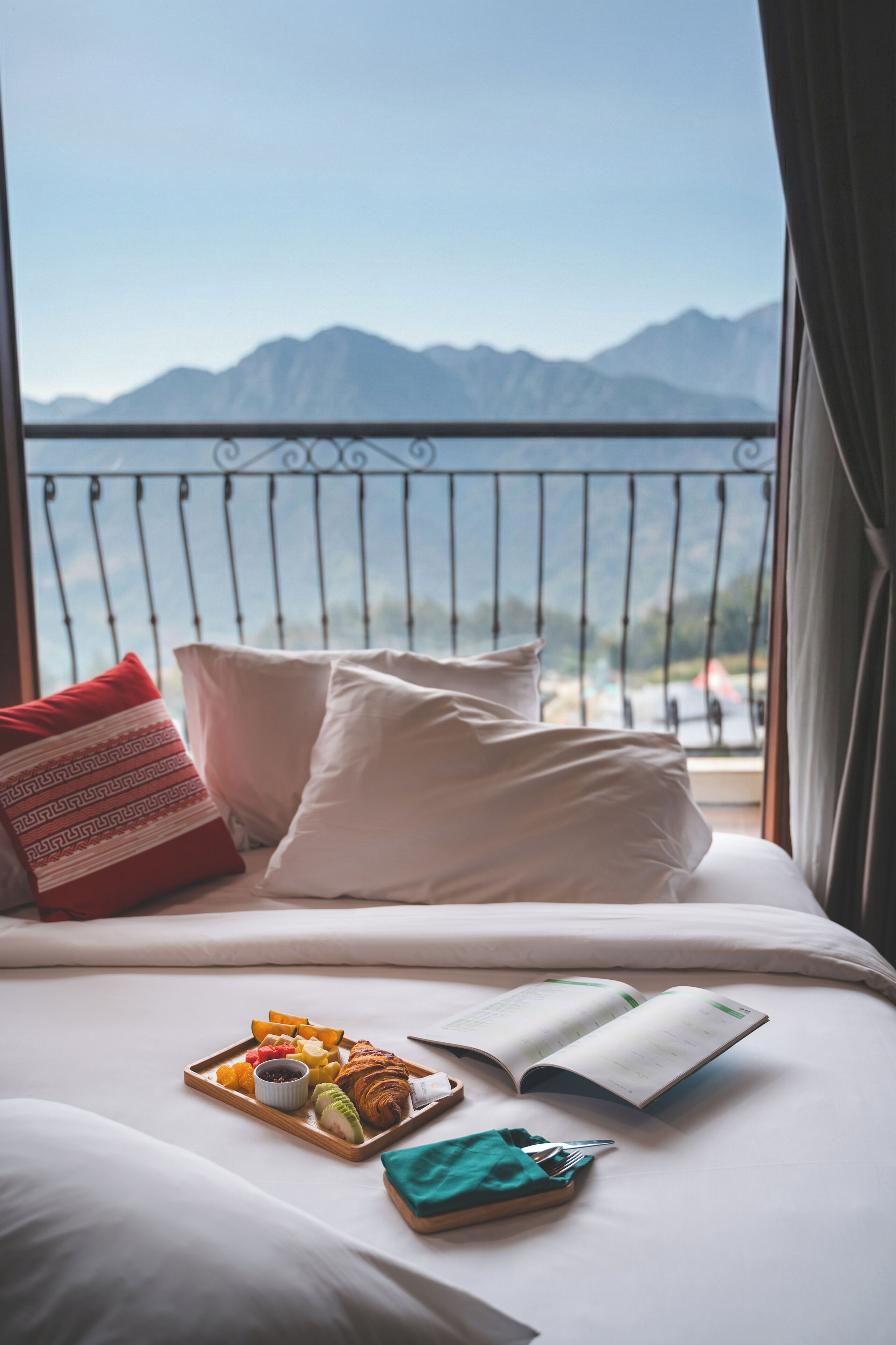 Una cama con una bandeja de desayuno, un libro y vistas a las montañas a través de la barandilla de un balcón.
