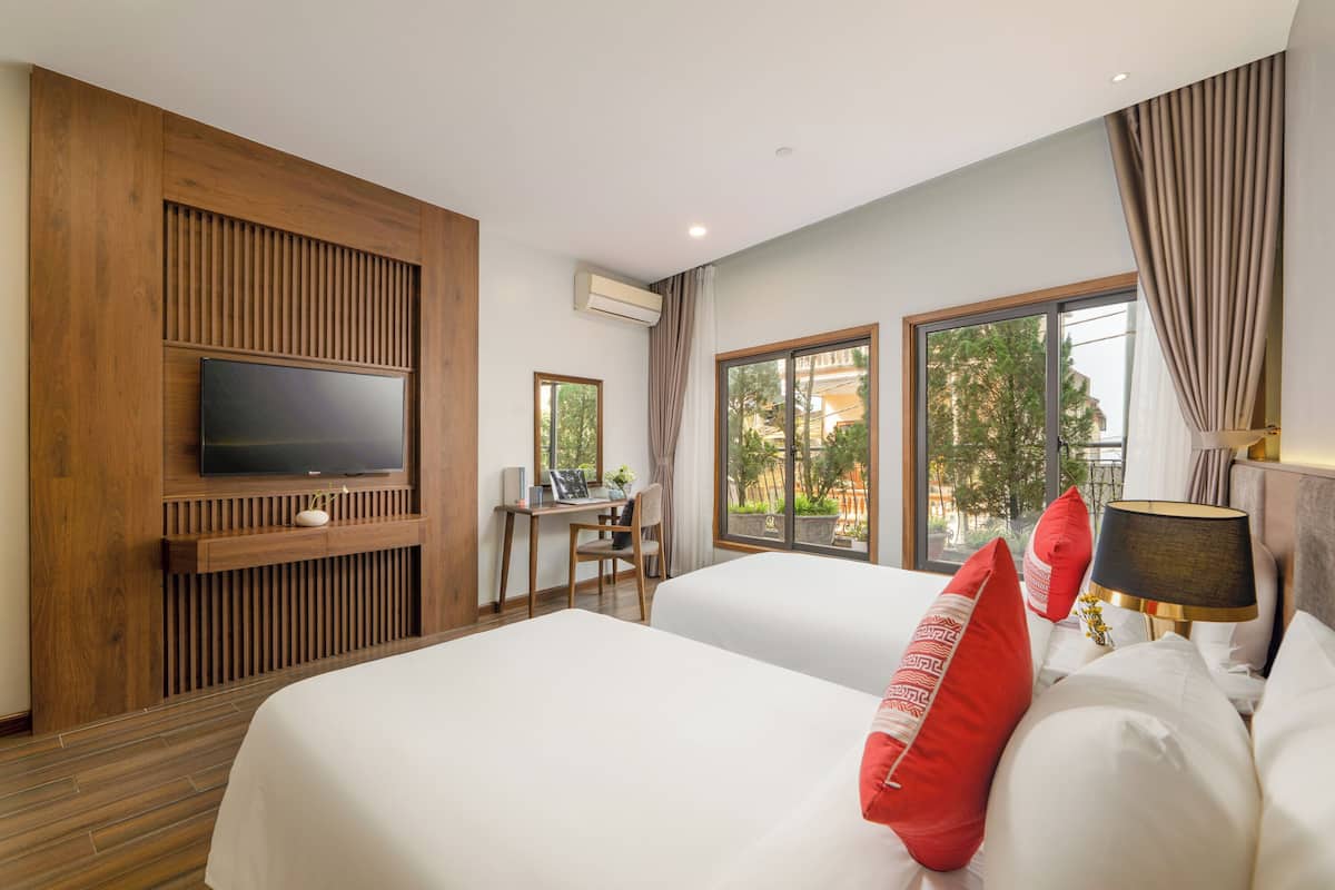 Habitación de hotel con dos camas, un televisor montado en un panel de madera, un escritorio con una silla y ventanas amplias con vistas a zonas verdes.