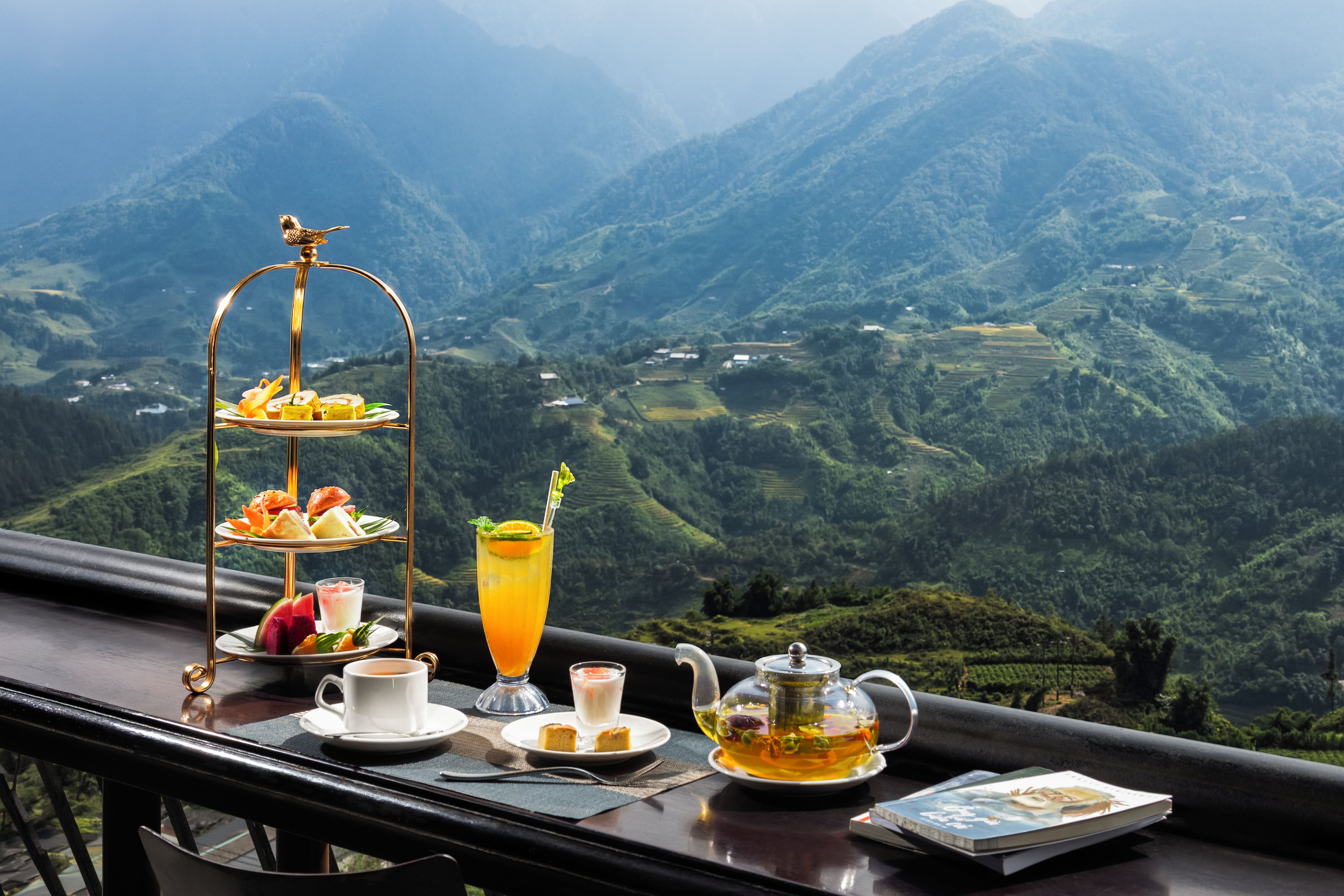 Una mesa con un juego de té, un vaso de zumo de naranja, una taza de café y una bandeja de pasteles, con vistas a la montaña.