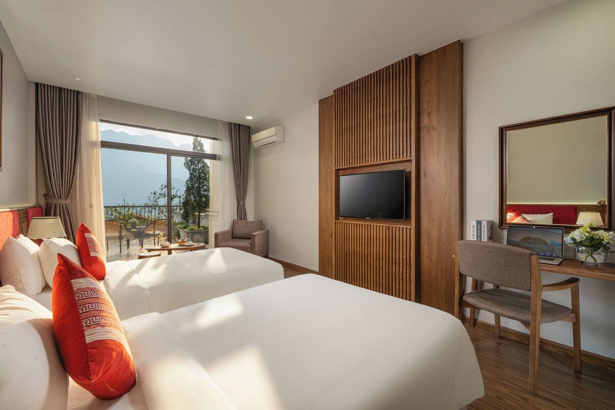 Habitación de hotel con dos camas, televisión, escritorio con silla y vista a las montañas desde la ventana.