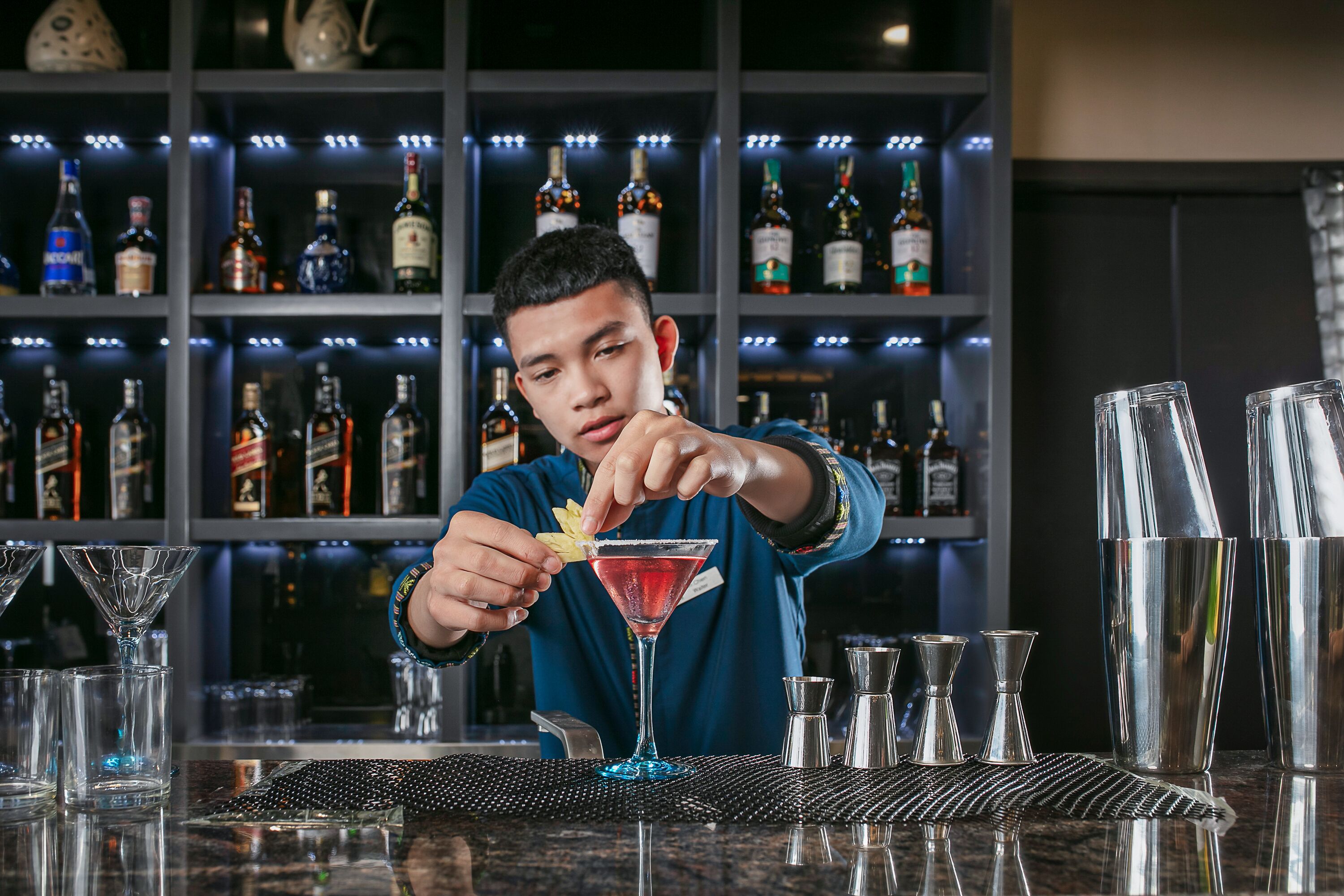 Un cantinero preparando un cóctel en un bar con estantes llenos de botellas de licor al fondo.