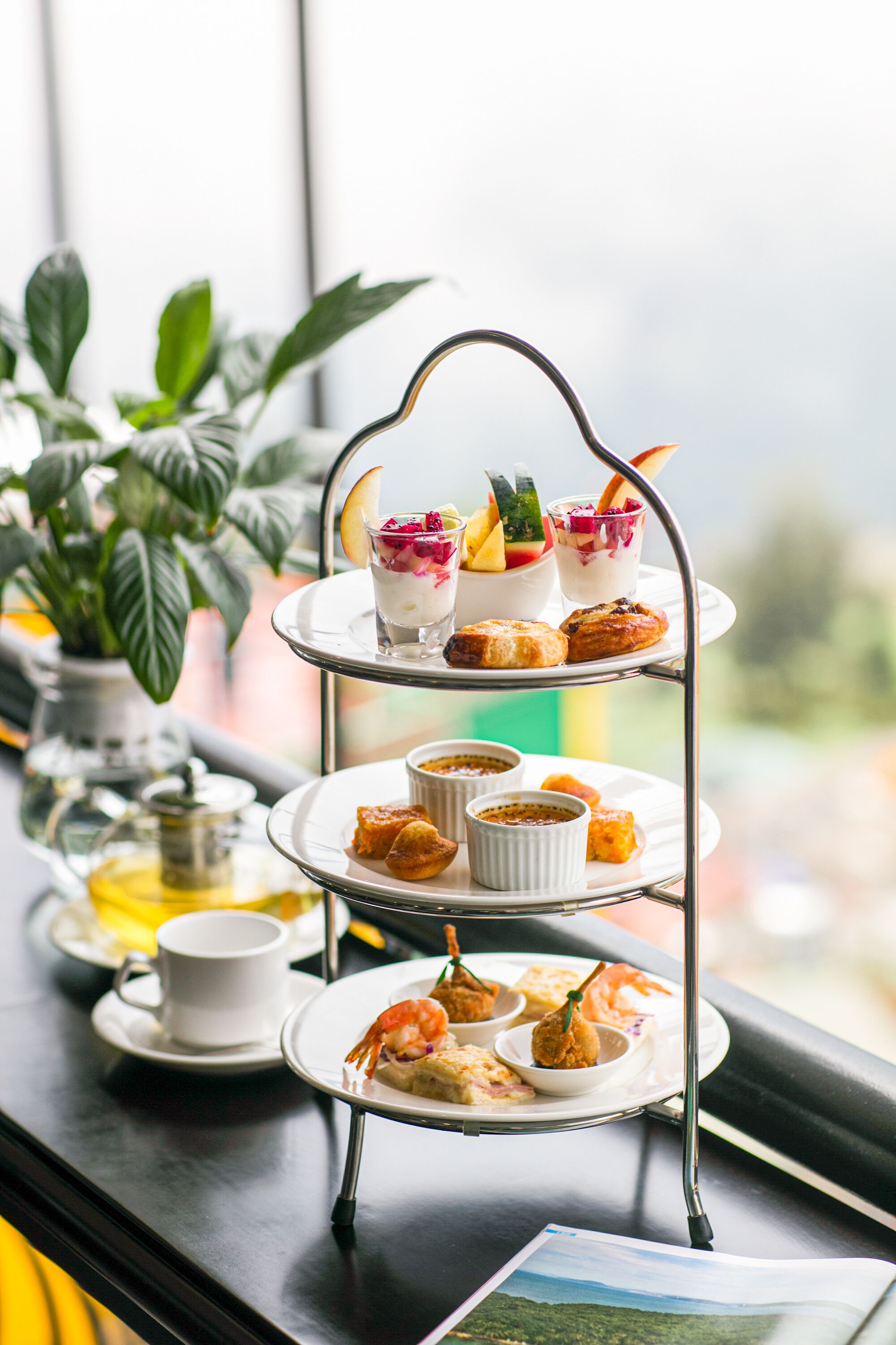 A tiered serving tray with various desserts and pastries, a cup of coffee, and a small potted plant on a table by a window.