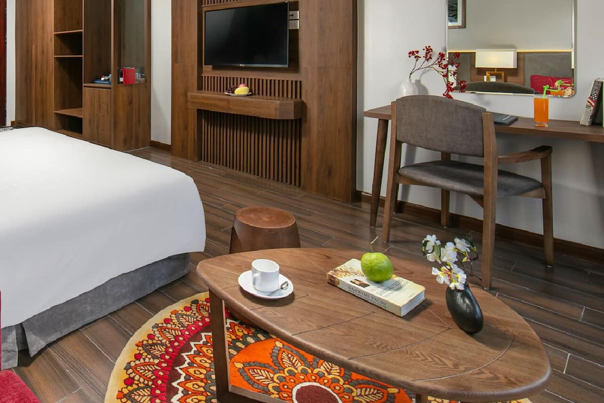 Habitación de hotel con cama, televisor montado en una pared de paneles de madera, escritorio con silla, una mesita con una taza y un libro, y una alfombra colorida.