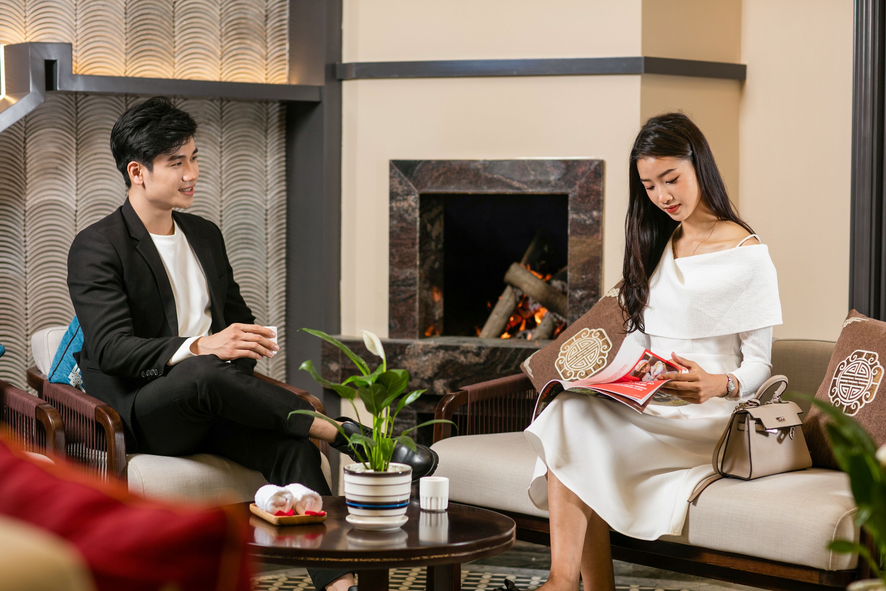 A man and woman sitting on a sofa in a well-lit room with a fireplace, a small table with a plant, and a decorative pillow.